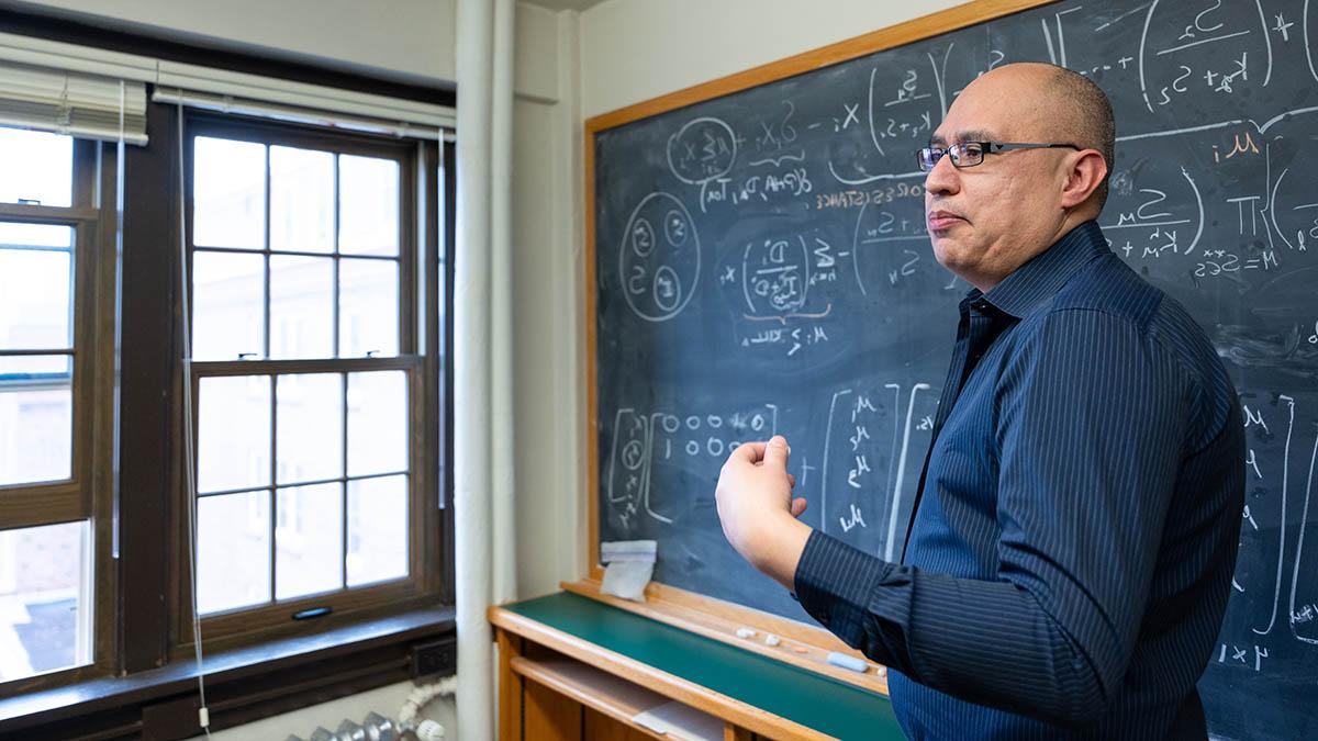 Man wearing glasses and blue shirt motions with his hand while standing in front of a mathematical formula on blackboard.