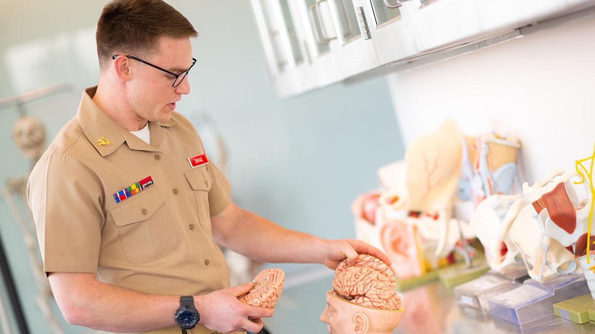 Young man wearing glasses and military blouse with ribbons and slacks stands in front of cupboard of anatomy models.