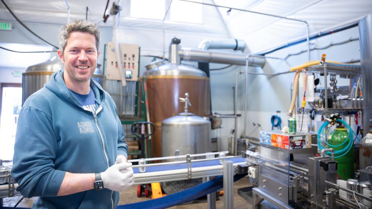 Man in blue sweatshirt stands in front of stainless steel equipment used to brew beer.